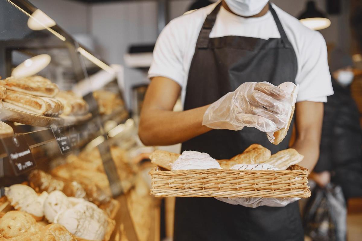 Employé en masque et tablier manipule des baguettes dans une boulangerie devant une vitrine de pains